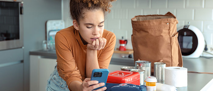 A person in a kitchen checks her hurricane preparedness guide on a smartphone. Nearby are supplies relevant to hurricane safety and hurricane season.